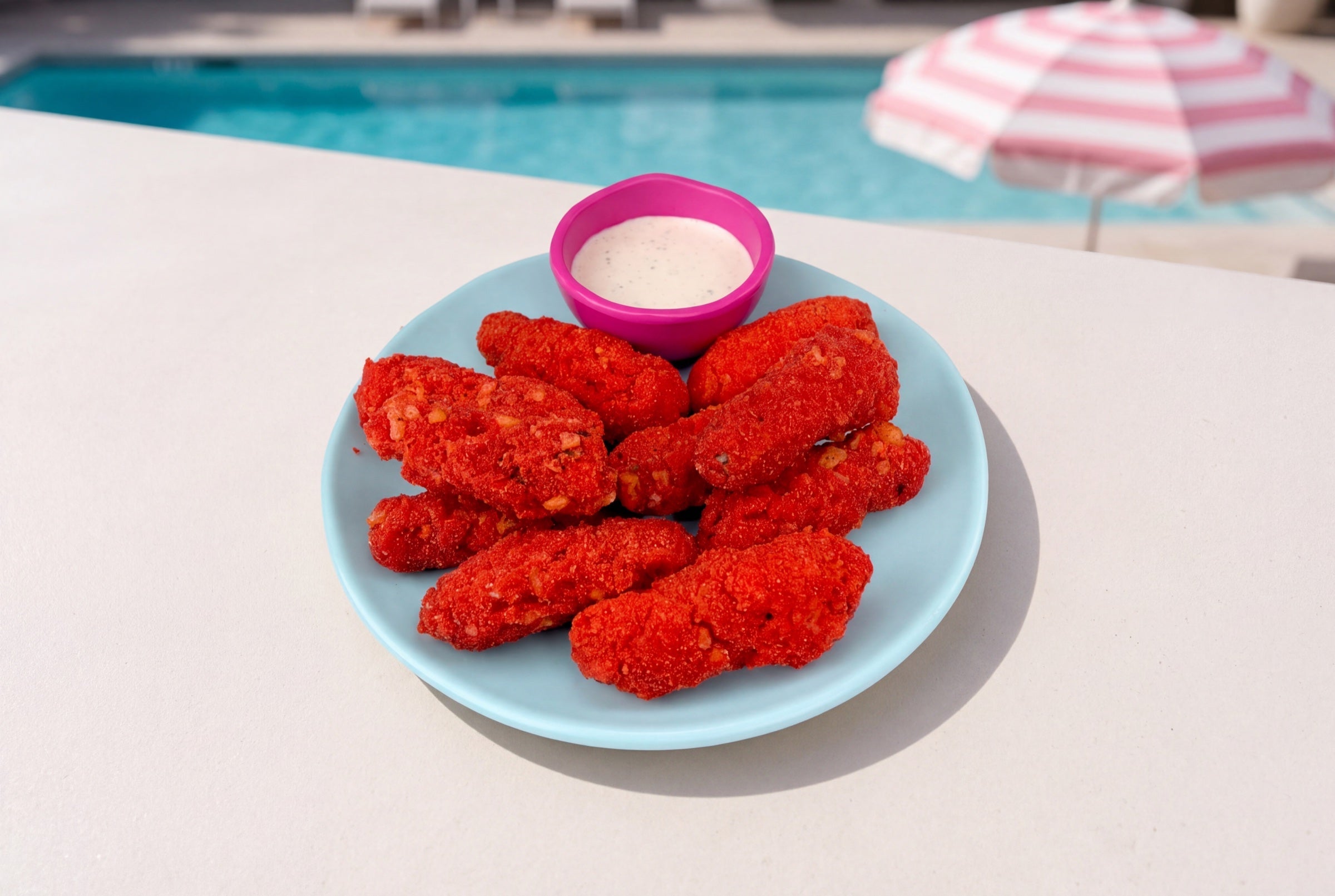 Bright red Flamin’ Hot tempura baby pickles with ranch on a light blue plate beside a sunny pool.