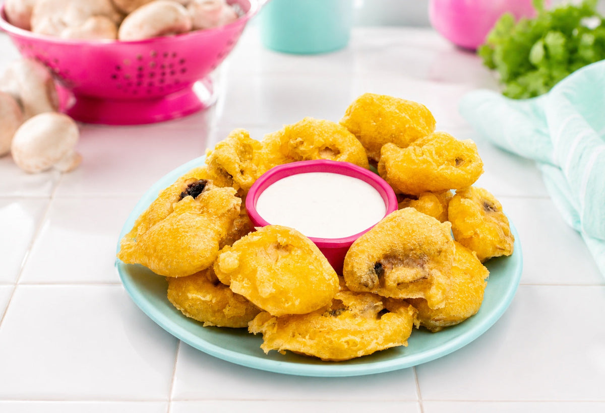 Tempura mushrooms plated with creamy dip on a white tile counter with fresh mushrooms in a pink colander